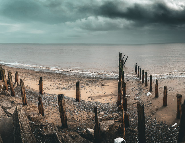 Broken down wooden sea defence posts gradually going into the North Sea. The sky is full of rain clouds and looks like it will rain soon. The beach is a mix of sand and stones.